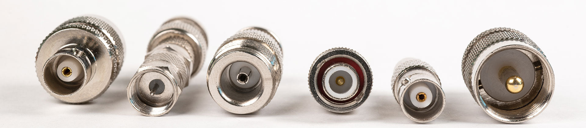 Close-up image of various coaxial cable connectors arranged side by side on a white background, displaying different connector types and their metallic finishes with detailed threading and central pins.