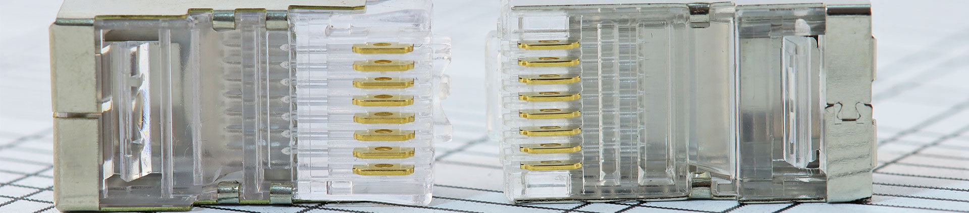Close-up image of two clear and metallic RJ45 Ethernet connectors placed side by side, showing detailed gold-plated pins and precision design on a graph paper background.