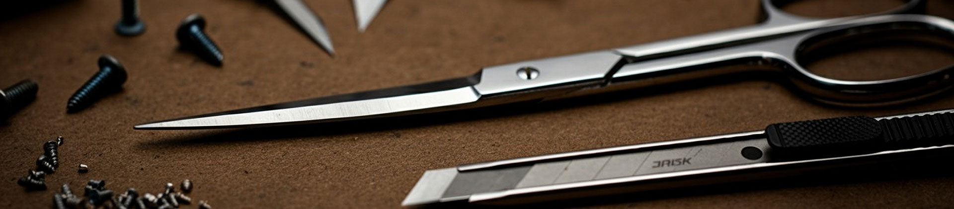 A close-up of precision tools, including stainless steel scissors and a utility knife, arranged on a textured brown surface with small screws scattered around.