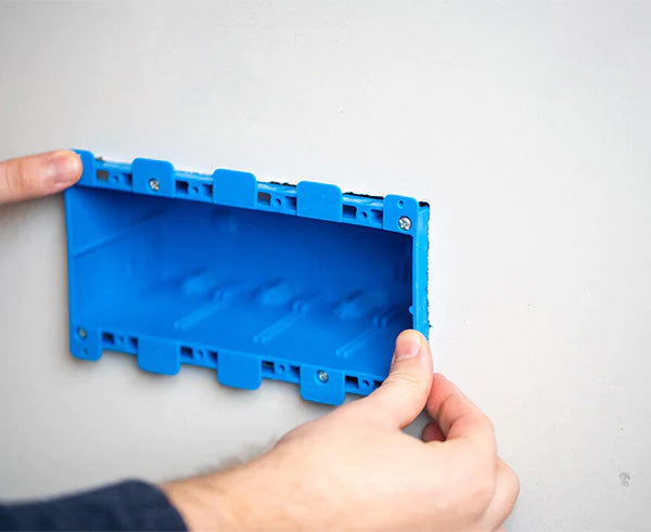 Inserting a blue non-metallic electrical box into a pre-cut opening in a wall using the template for guidance